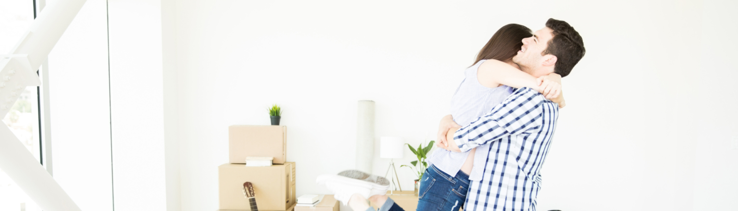 Junges Paar umarmt sich im neuen zu Hause. Im Hintergrund sind Umzugskartons zu sehen