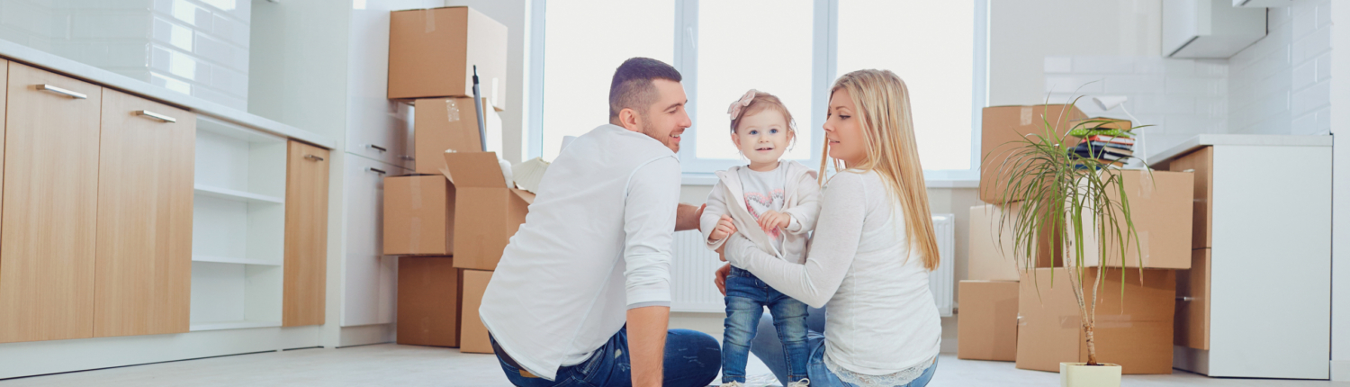 Eine junge Familie mit einer kleinen Tochter sitzt auf dem Fußboden der Küche im neuen zu Hause, um Sie herum sind Umzugskartons - Martina Wagner Immobilien
