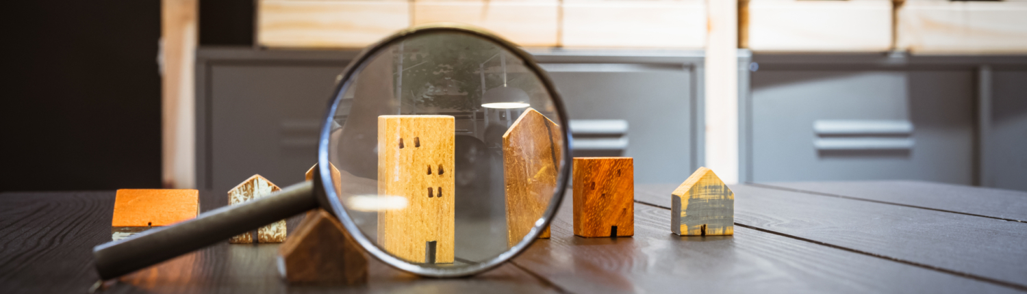 Haus gesucht Verschieden Hausmodelle aus Holz stehen auf einem Holztisch. Eine Lupe vergrößert Sie - Martina Wagner Immobilien