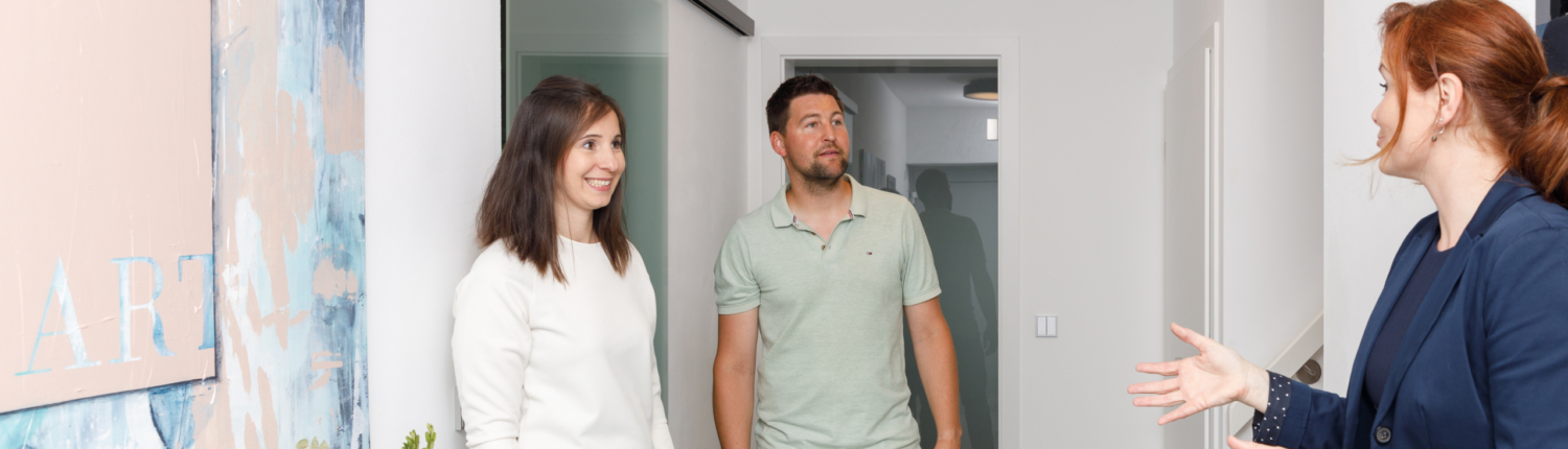 Besichtigungstermin. Martina Wagner erklärt einem jungen Paar etwas im Flur eines modernen Hauses. - Martina Wagner Immobilien