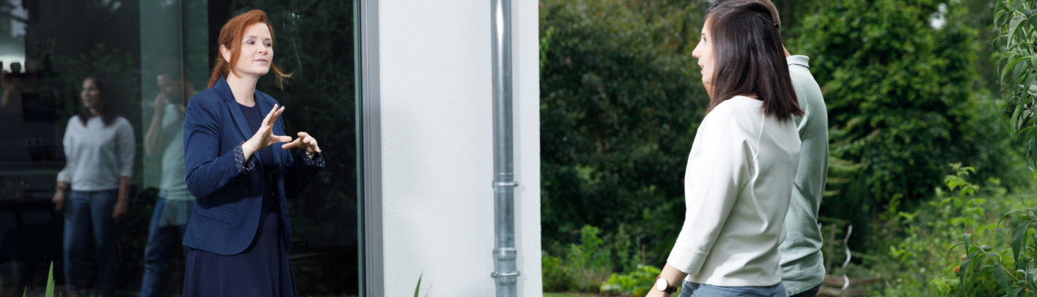 Besichtigungstermin. Martina Wagner erklärt einem jungen Paar etwas im Garten, im Hintergrund befindet sich die Immobilie - Martina Wagner Immobilien