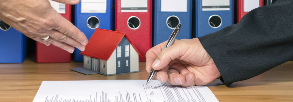 Maklervertrag. Ein Mann unterschreibt einen Vertrag der auf einem Tisch liegt im Hintergund befinden sich Aktenordner und die Miniatur einer Immobilie. Ein anderer Mann deutet auf den Vertrag - Martina Wagner Immobilien