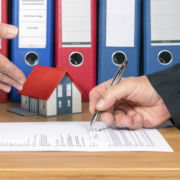 Maklervertrag. Ein Mann unterschreibt einen Vertrag der auf einem Tisch liegt im Hintergund befinden sich Aktenordner und die Miniatur einer Immobilie. Ein anderer Mann deutet auf den Vertrag - Martina Wagner Immobilien