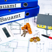Auf einem Grundriss liegen Bauamtordner, ein Taschenrechner, eine Hausminiatur, Schlüssel, Geldstücke, Minibausteine und ein Kugelschreiber - Martina Wagner Immobilien