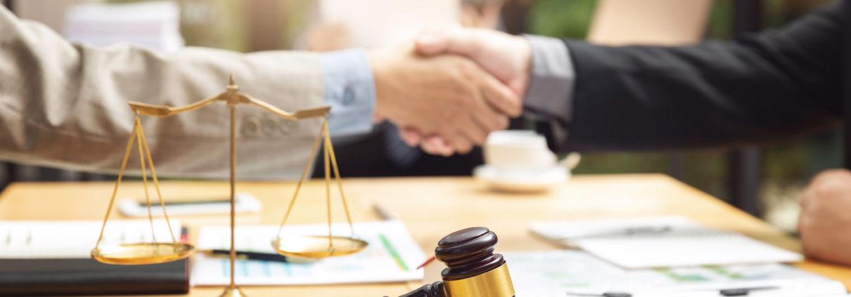 Ein Handschlag besiegelt einen Vertrag im Vordergrund sieht man eine Waage sowie einen Richterhammer und Dokumente - Martina Wagner Immobilien