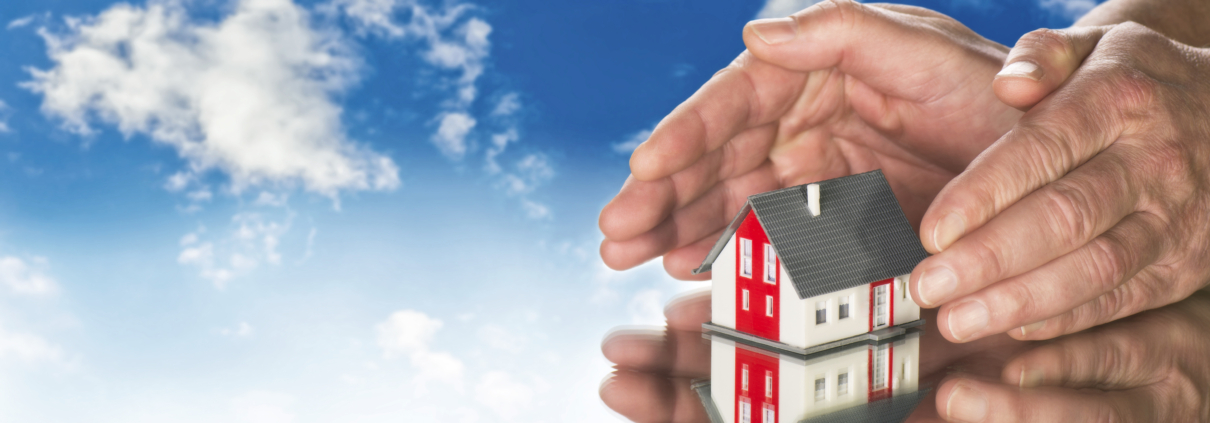 Eine Hausminiatur steht auf einem Spiegel und wird von 2 Händen beschirmt, darüber sieht man einen blauen Himmel mit Wolken - Martina Wagner Immobilien