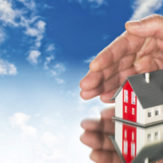 Eine Hausminiatur steht auf einem Spiegel und wird von 2 Händen beschirmt, darüber sieht man einen blauen Himmel mit Wolken - Martina Wagner Immobilien