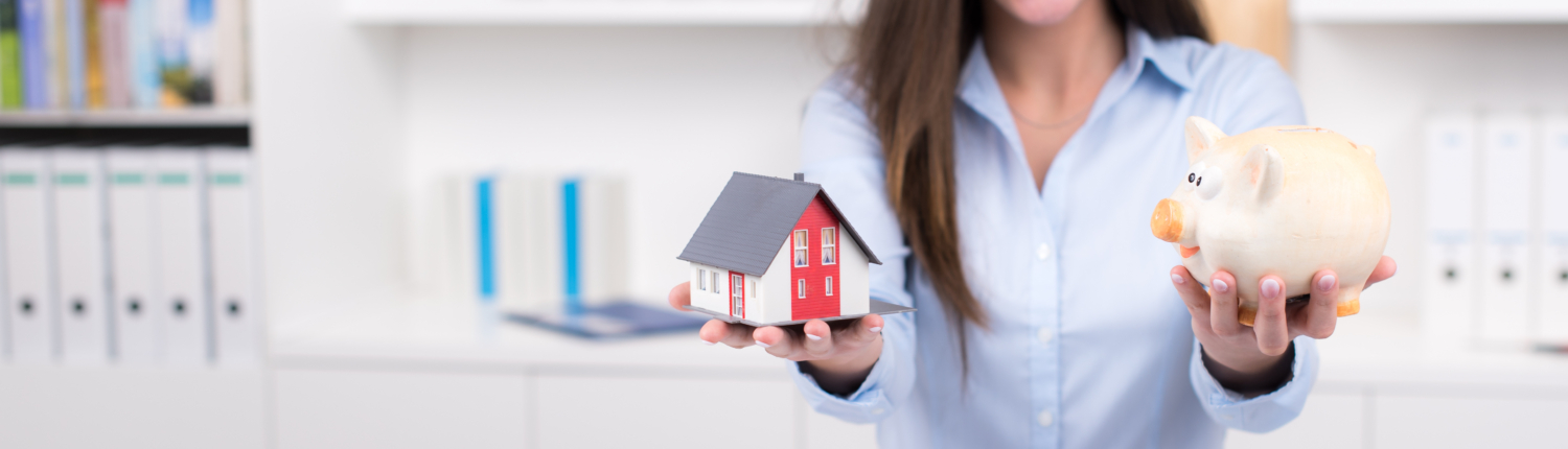 Eine Maklerin hält in der einen Hand das Modell eines Einfamilienhauses und in der anderen ein Sparschwein