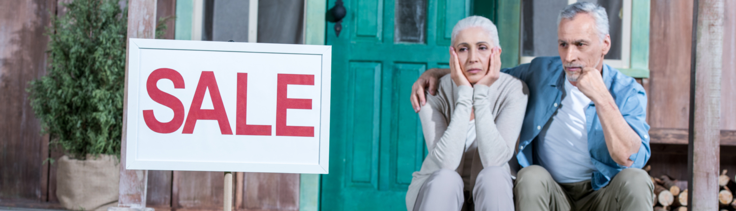 älteres Ehepaar sitzt auf der Treppe vor einem Haus, links neben steht ein "zu Verkaufen Schild".