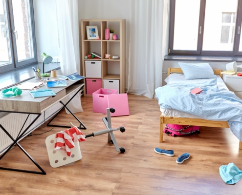 Man sieht einen Schreibtisch links und ein Bett rechts in einem unaufgeräumten Jugendzimmer , der Drehstuhl ist umgekippt, Dinge liegen auf dem Boden.