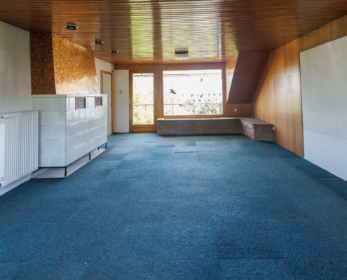 Blick auf ein großes Wohnzimmer mit Kachelofen links, großer Fensterfront geradeaus. Blauer Teppich, Holzdecke und teilweise Holzvertäfelung.