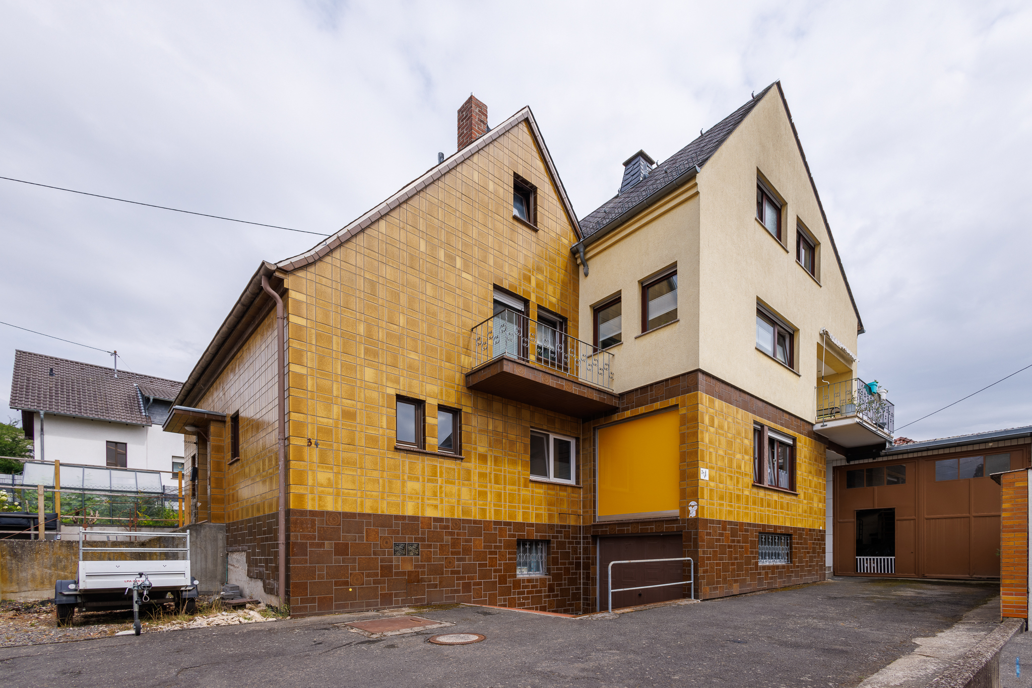 Außenansicht Immobilie von der Seite, Doppelhaus in den Farben braun, ocker und gelb. Teilweise gefliest, teilweise verputzt, rechts braunes Tor zur Halle, links steht ein Anhänger im Vordergrund Einfahrt.