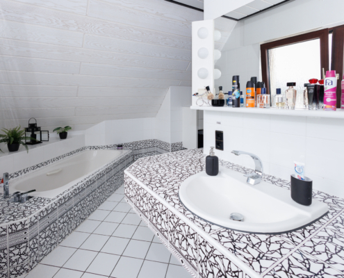 Waschbeckenbereich des Bad en suite Badezimmer in schwarz weiß gehalten. Gemusterte Fliesen um das Waschbecken rechts, darüber ein Spiegel mit Abstellfläche und diversen Produkten, im Spiegel sieht man das Fenster. Links Eckbadewanne mit denselben Fliesen, auf Ablage stehen 2 Pflanzen und eine schwarze Laterne, dahinter beginnt Dachschräge.