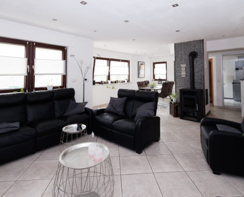 moderner Wohn-Essbereich mit vielen Fenstern Wohn-Essbereich, heller Fliesenboden, drei Fenster links und hinten, davor schwarzer Couchen und silberne Coffeetables, rechts schwarzer Sessel, dahinter schwarzer Kaminofen, dahinter rechts Glasfront und Küchenzeile in grau zu sehen, links Essbereich mit Tisch und 4 Stühlen.