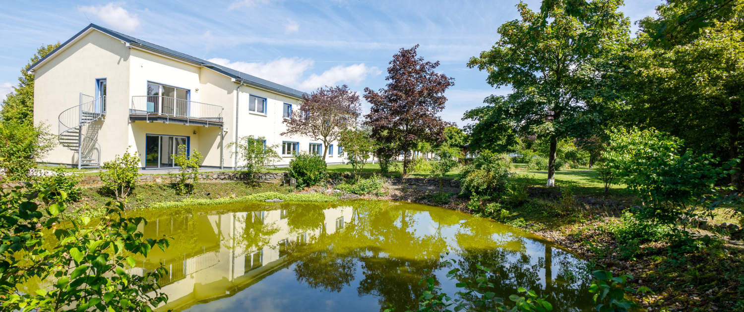 Blick über einen großen Teich mit verschiedenen Laubbäumen umsäumt, im Hintergrund Immobilie gelb verputzt, 2 stöckig, im Voderbereich Wendeltreppe und Balkon, blauer Himmel