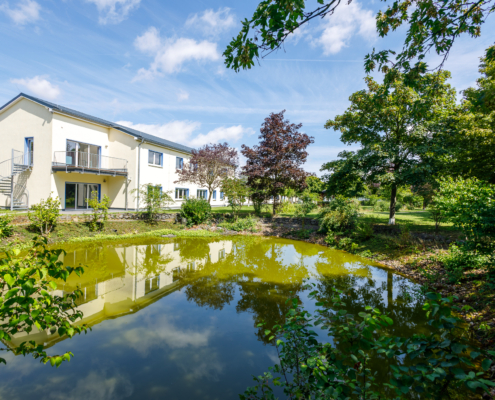 Blick über den Löschteich auf die Gewerbeimmobilie Blick über einen großen Teich mit verschiedenen Laubbäumen umsäumt, im Hintergrund Immobilie gelb verputzt, 2 stöckig, im Voderbereich Wendeltreppe und Balkon, blauer Himmel