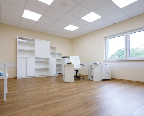 Mayen Göth Büro 1. Etage großer Büroraum im 1. OG, Holzfußboden, weißes Mobiliar (Regalwand, Schreibtisch mit weißem Drehstuhl), rechts Fenster