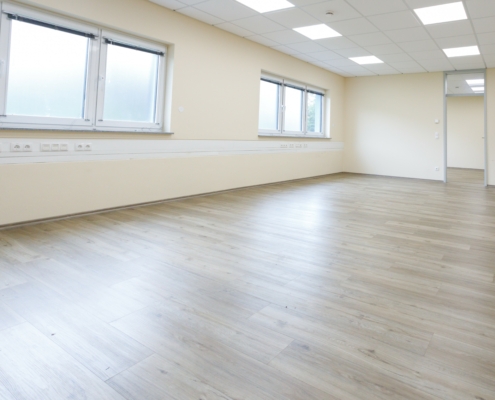 Mayen Großraumbüro Großraumbüro, Fensterfront links darunter IT Leiste, Boden heller Holzboden, im hinteren Bereich Zugang zu anderem Raum