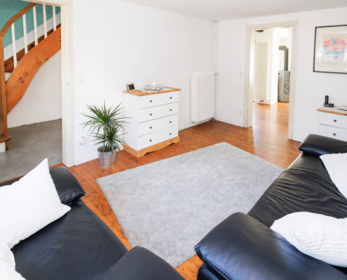 Wohnzimmer mit Blick in den Flur und den Essbereich, man sieht die Couchmöbel, Leder schwarz mit weißen Kissen im Anschnitt, davor grauer Teppich und weiße Kommode, Holzdielenboden, rechts Türöffnung zum Esszimmer, man sieht den Kamin, links Öffnung zum Flur, man sieht die Holztreppe.