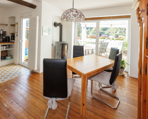 Blick über den Essbereich in die Küche und nach draußen. Links Küchenzeile und Fliesenboden mit Terrassentür, rechts Holzschrank davor Tisch mit 4 Stühlen, Echtholzdielen, Specksteinofen und Terrassentür.