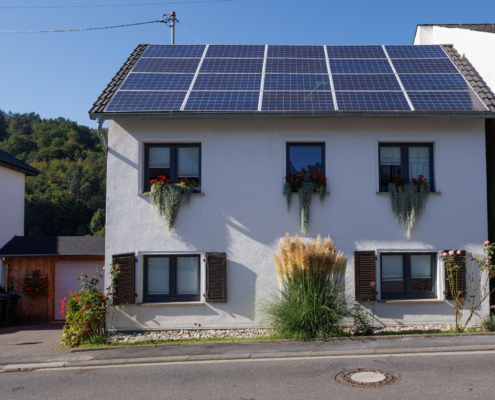 Frontansicht Haus, weiß verputzt, PV Anlage auf Dach, Fenster im OG haben Blumekästen mit hängenden Pflanzen, Fenster unten haben Fensterläden in Braun. frontal Pampasgras, links Garage aus Holz mit weißen Tor, daneben Mülltonnen, im Bildvordergrund Straße