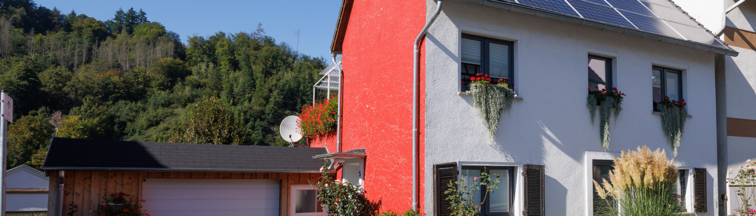 Frontansicht Immobilie, Front weiß, Dach mit PV Anlage, linke Seite rot verputzt, daneben Holzgarage mit weißem Tor und weißer Tür. Im Hintergrund Wald,