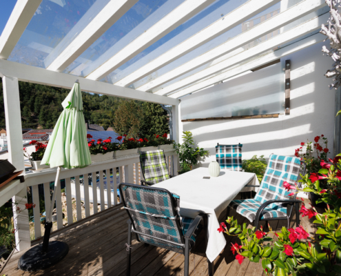 Terrasse mit Holzboden und weißer Überdachung, Tisch mit 4 Stühlen. Pflanze im Vordergrund