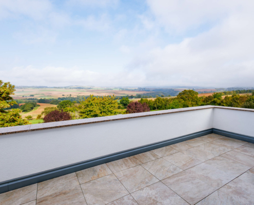 große Dachterrasse mit modernen Fliesen und halbhoher gemauerter (weiß verputzter) Brüstung, dahinter Blick aufs Maifeld mit blauem Himmel mit Schleierwolken