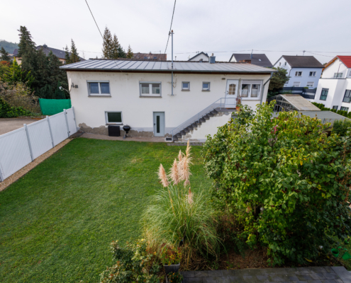 Blick aus dem OG in den Garten Erhöhter Blick über den Garten mit Begrünung rechts und Wiese auf das Nebengebäude, weißer Zaun links, Häuser im Hintergrund.