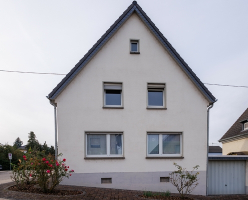 Seitenansicht Einfamilienhaus mit Garage Seitenansicht Immobilie, Spitzdach, im unteren Bereich 2 große Fenster, darüber 2 kleinere Fenster, darüber mittig ein kleines Fenster, rechts neben dem Haus eine Garage mit grauem Tor, Haus weiß verputzt mit hellgrauem Sockel. Davor Rosensträucher