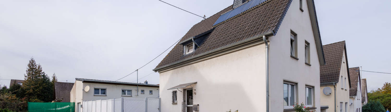Ansicht Grundstück mit EFH mit 4 Solarplatten auf dem Dach, Eckgrundstück im Vorderbereich Gehweg und Pflaster mit Rosensträuchern. Neben dem Gebäude ist ein weißer Zaun zu sehen, dahinter Gebäude mit Flachdach und Bäume und Zaun.