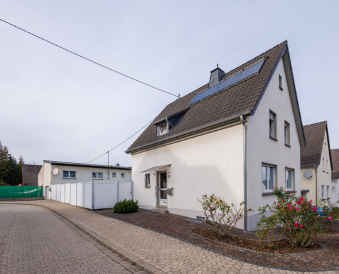 Titelbild Ansicht Grundstück mit EFH mit 4 Solarplatten auf dem Dach, Eckgrundstück im Vorderbereich Gehweg und Pflaster mit Rosensträuchern. Neben dem Gebäude ist ein weißer Zaun zu sehen, dahinter Gebäude mit Flachdach und Bäume und Zaun.