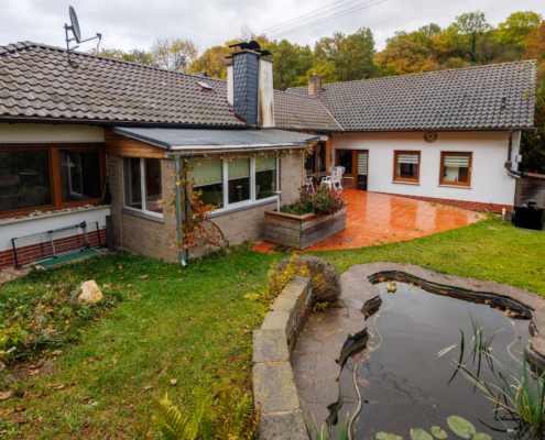 Blick auf ein Haus in Winkelbauweise mit halbrunder Terrakottafliesenterrasse, im Vordergrund rechts Gartenteich, links Wiese.