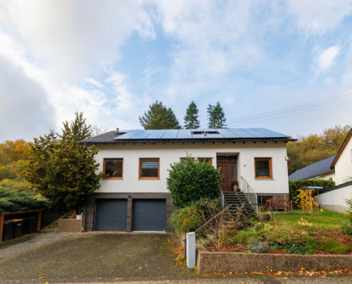 Außenansicht Immobilie, Links Abstellschuppem mit Mülltonnen, Mittig 2 Garagentore und rechts Treppenaufgang zur Eingangtür, daneben Vorgartenbereich mit Tanne. Das Dach hat eine PV Anlage, der Himmel ist blau mit Schleierwolken
