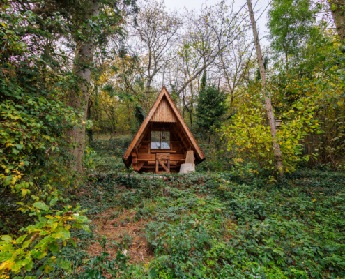 Vordergrund grüner Waldbodenbewuchs in leichter Handlage, Im Hintergrund und an den Seiten Laubbäume. Mittig eine Hütte, mit sehr spitzwinkligem Dach, Sitzgelegenheit, Tisch und Fenster