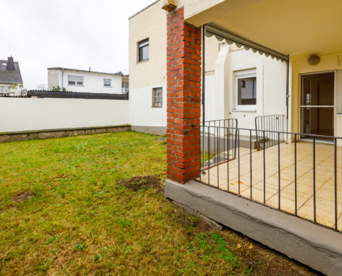 Blick in den Garten Man sieht links eine überdachte Terrasse mit Brüstung, links und im Hintergrund eine Wiese. Rechts das Haus.
