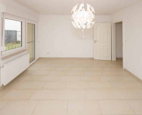 Wohnbereich mit Zugang zu Terrasse und Garten Raum mit hellen Fliesen, links ist ein Fenster und eine Terrassentür, rechts eine Zimmertür. Mittig hängt eine weiße Lampe.