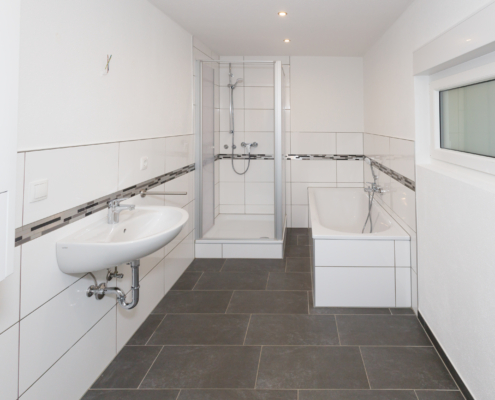 modernes Tageslichtbad mit Wanne und Dusche Badezimmer mit grauen und weißen Fliesen, rechts ist ein kleines Fenster und die Wanne. Links ist im Hintergrund die Dusche und vorne das Waschbecken.