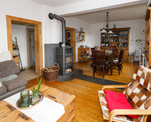 Blick über den Wohnbereich mit Couch, Couchtisch und Schwingsessel in den Essbereich mit Esstisch, Buffetschrank und verschiedenen Regalen rechts. Links sieht man noch einen Kaminofen und die beiden Türöffnungen