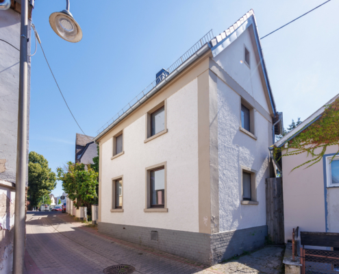 Außenansicht Außenansicht einer Altbauimmobilie mit Straße im Vordergrund und blauem Himmel. Das aus hat ein spitzes Satteldach, einen gräulichen Klinkersockel und ist hell verputzt, die Außenseiten und Abgrenzung zum Dachstuhl sind etwas dunkler gestrichen