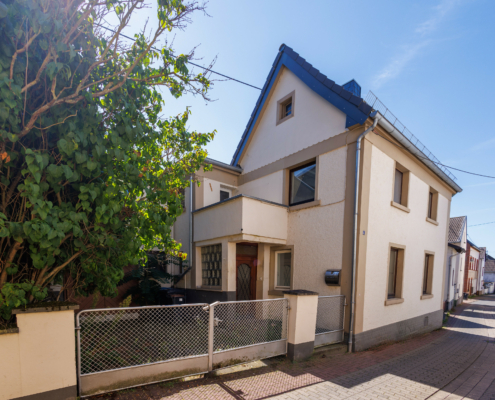 Seitenansicht Immobilie Seitenansicht einer Altbauimmobilie, strahlend blauer Himmel eine leicht kurvige gepflasterte Straße rechts, die Immobilie links, davor ein großer Baum