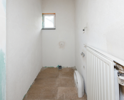 Gäste WC, Wände weiß, sehr schmal, hinten kleines weißes Fenster, rechts Heizung, Boden modern gefliest, ansonsten nur Rohre, Waschbecken steht auf Boden rechts.