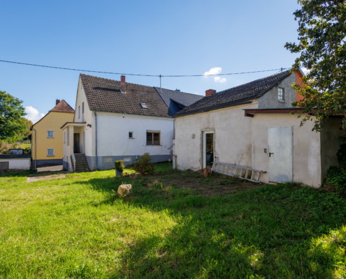 Blick über eine grüne Wiese, am rechten Bildrand ist ein Laubbaum davor 2 Nebengebäude und davor die Doppelhaushälfte, blauer Himmel