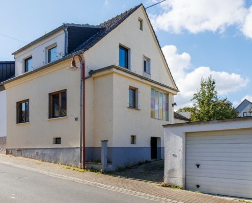 Seitenansicht Doppelhaushälfte, man sieht rechts am Bildrand eine Garage, daneben eine Einfahrt, dann gelblich verputzte DHH mit grauem Sockel, davor rote Straßenlaterne und Straße. Himmel blau leichte Wolken