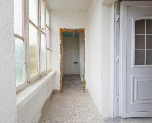 Windfang mit Bleiverglasung links, dahinter erkennt man eine Öffnung zu einem schmalen Raum, davor schmaler Flur, rechts öffnet sich die weiße Haustür