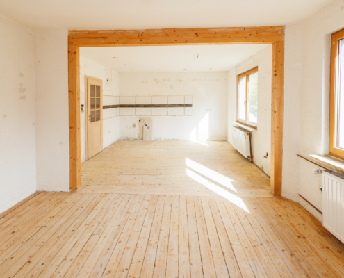 Wohn-Ess-Küchenbereich mit Echtholzdielen, rechts 2 große Fenster, darunter Heizkörper, mittig befindet sich ein Bogen aus Nadelholz, hinten ein Fliesenspiegel an der Wand, Zimmertür im Landhausstil