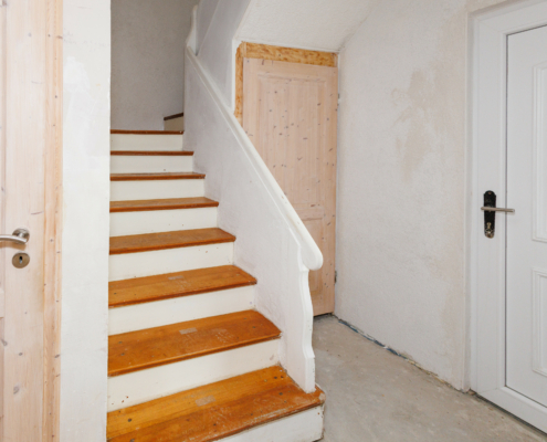 Eingangsbereich eines Hauses, Zentral sieht man eine Altbauholztreppe, weißen Handlauf, Stufenseiten weiß, Tritte holzfarben, rechts Hauseingangstür weiß, daneben, Zugang zum Keller, links neben der Treppe sieht man noch eine Zimmertür