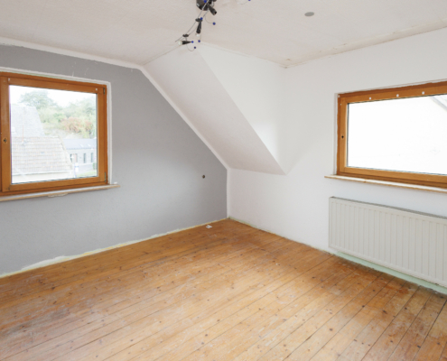 Zimmer mit leichter Dachschräge links, die linke Wand ist grau gestrichen und hat ein großes Holzfenster, rechte Wand weiß ebenfalls mit großem Fenster und Heizkörper darunter, Decke weiß, Fußboden Echtholzdielen.