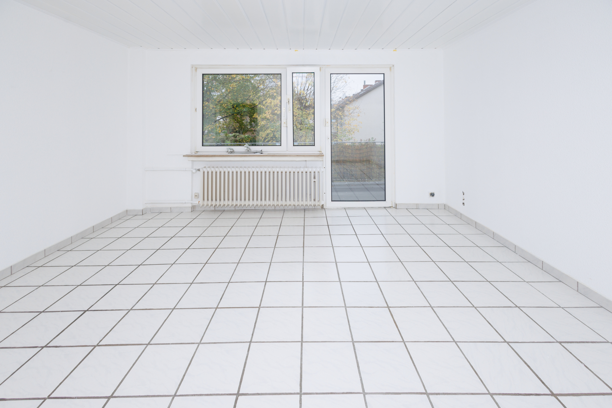 Wohnzimmer mit weißen Fliesen, Wänden und Decke, zentral ein Fenster mit Balkontür darunter ein Heizkörper