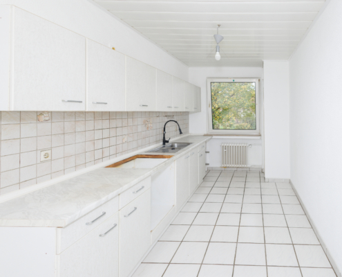 große Küche mit zeitlosem Mobiliar lange schmale Küche, weiße Fliesen, weiße Einbauzeile über die gesamte linke Bildhälfte, hinten Fenster, Decken und Wand ebenfalls weiß